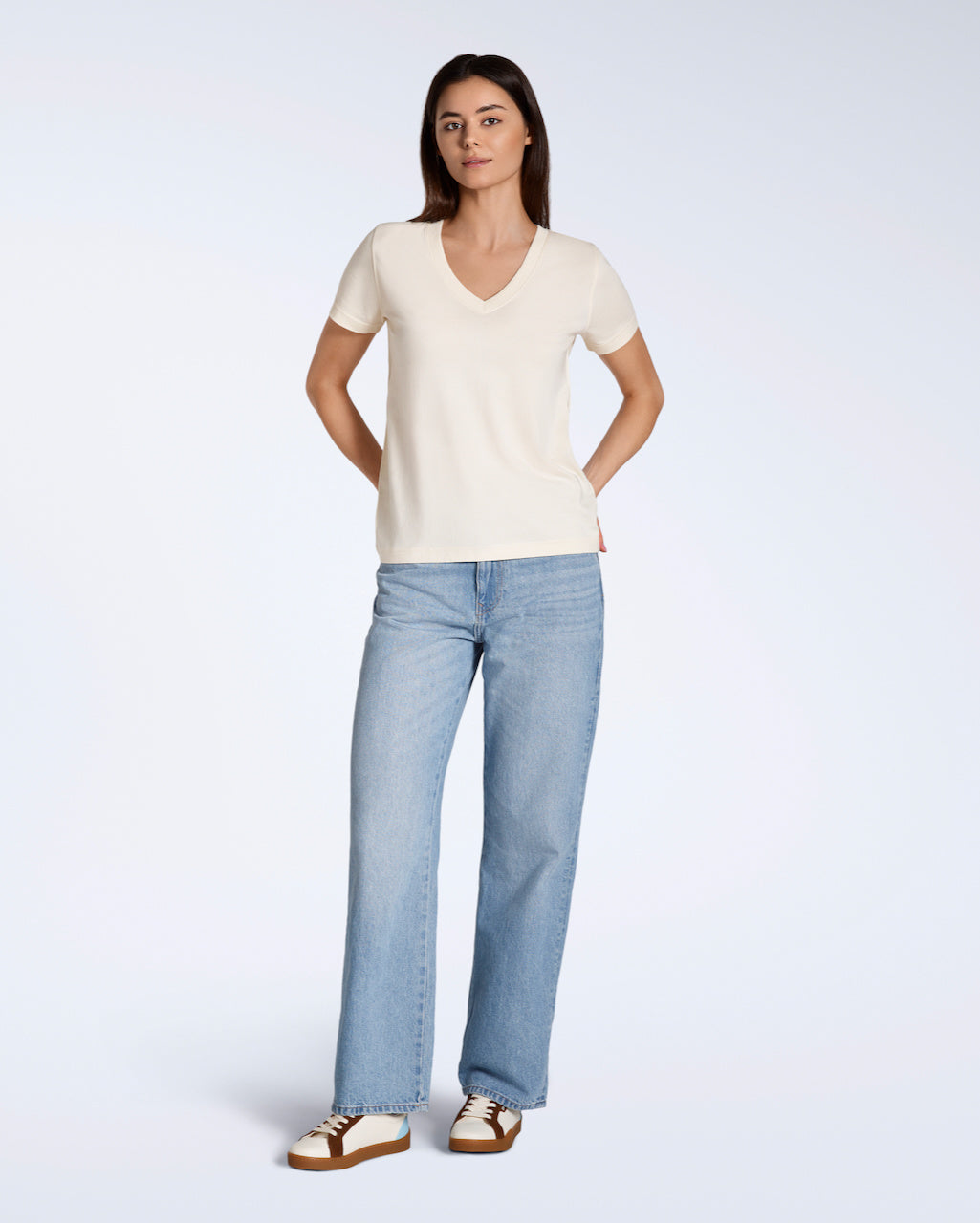 A full body photo of a woman looking into the camera with her hands by her hips, modelling a plain natural coloured, organic cotton v neck t shirt with a pair of blue jeans.