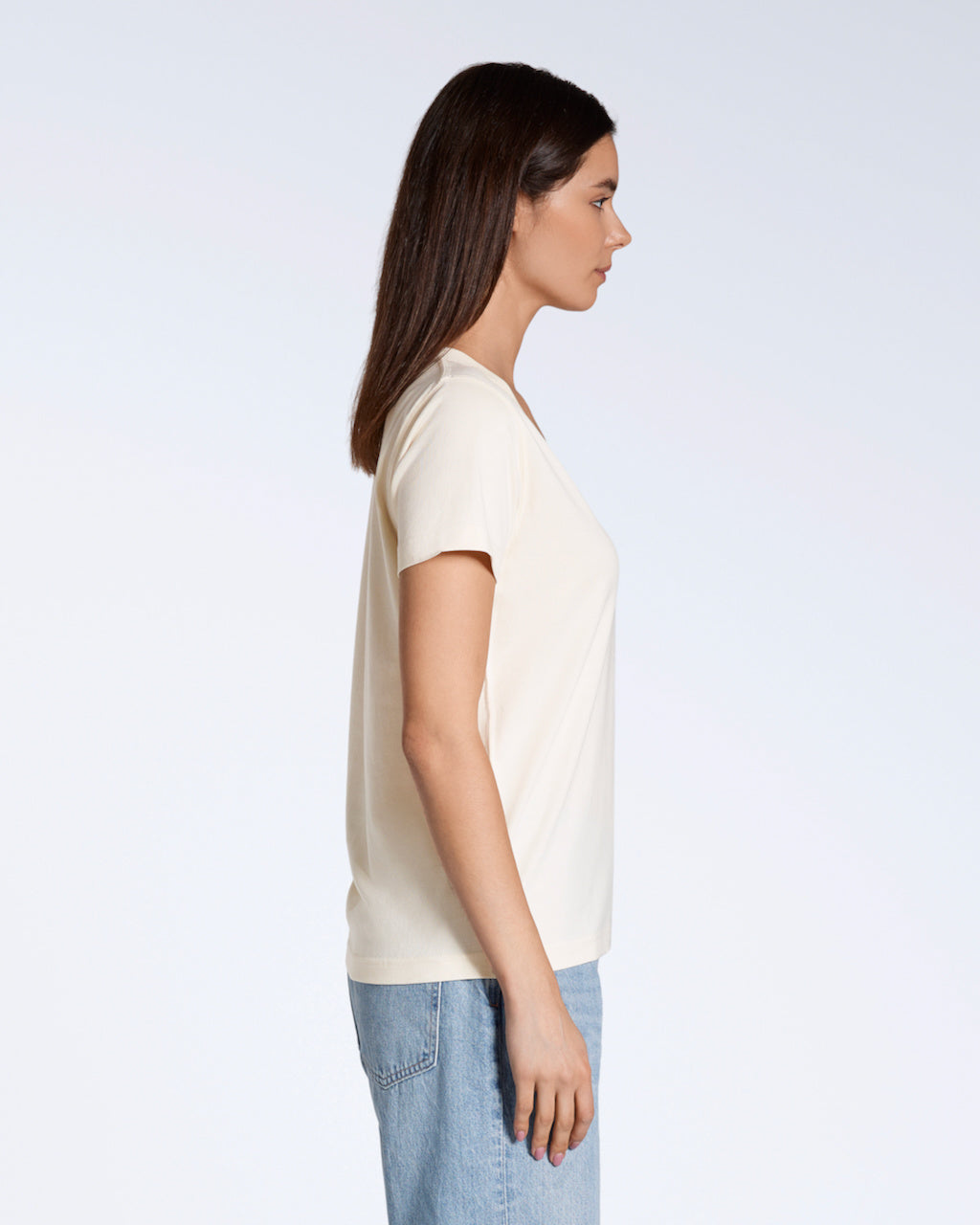 A woman facing side on, with her arms by her side, modelling a plain natural coloured, organic cotton v neck t shirt.