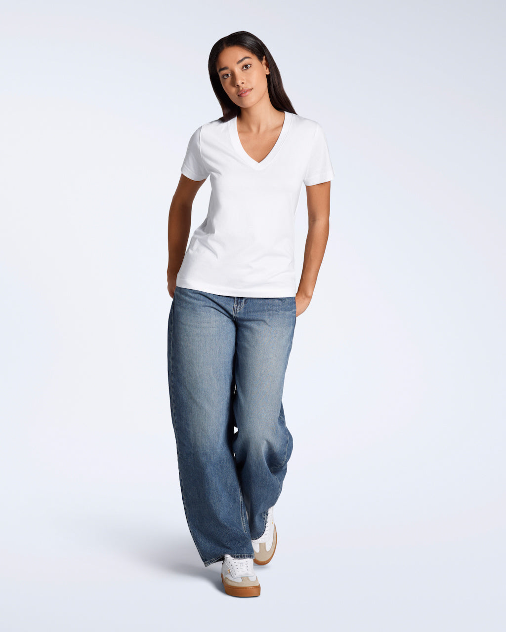 A full body photo of a woman looking into the camera, modelling a plain white, organic cotton v neck t shirt with a pair of blue jeans.
