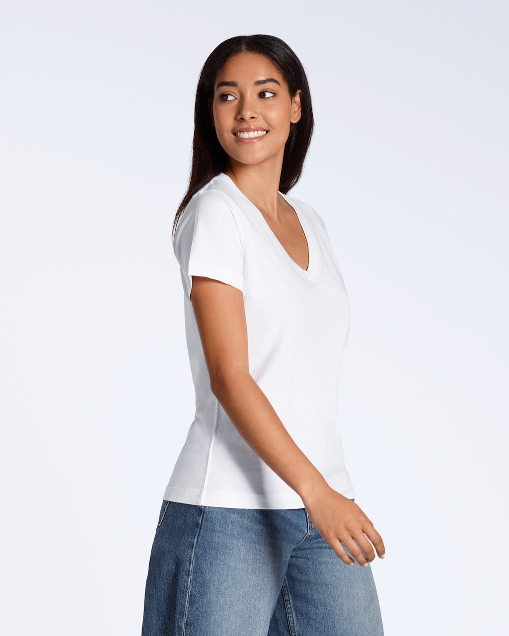 A woman smiling looking away from the camera, modelling a plain white, organic cotton v neck t shirt with a pair of blue jeans.