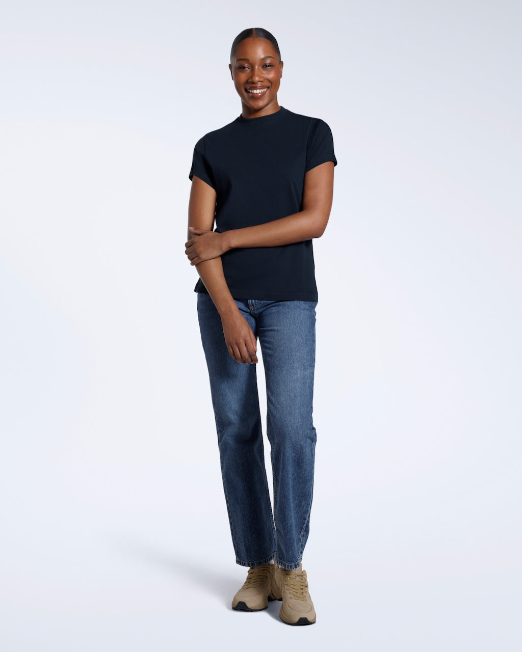 A full body photo of a woman smiling touching her arm, modelling a plain navy short sleeve organic cotton t shirt.