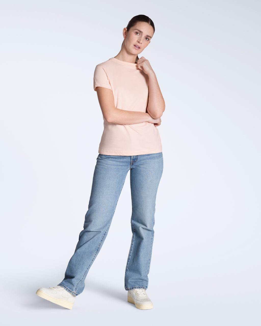 A full body photo of a woman looking into the camera, modelling a plain pink, short sleeve organic cotton t shirt with a pair of jeans.