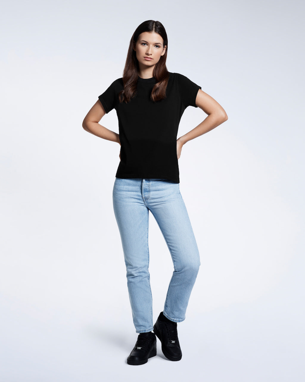 A full body photo of a woman with her hands by her waist, modelling a plain black short sleeve organic cotton t shirt.