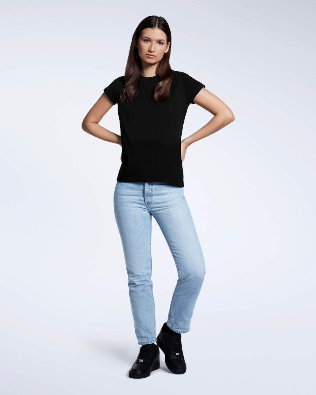 A full body photo of a woman with her hands by her waist, modelling a plain black short sleeve organic cotton t shirt.