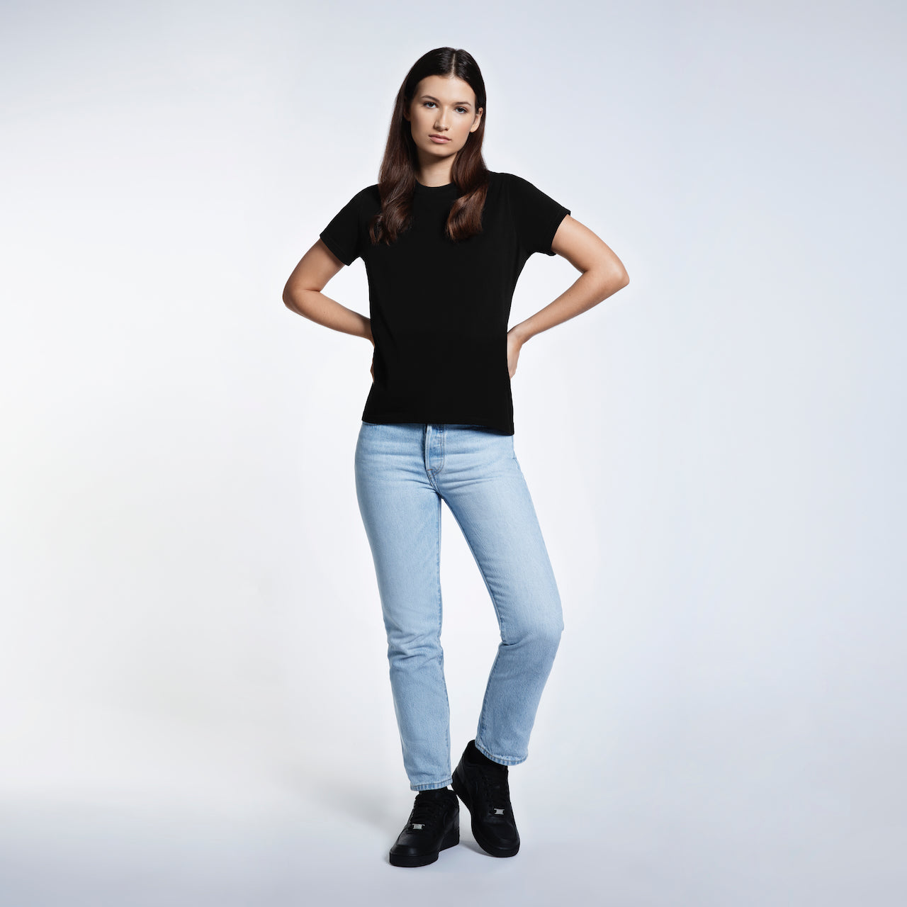 A full body photo of a woman with her hands by her waist, modelling a plain black short sleeve organic cotton t shirt.