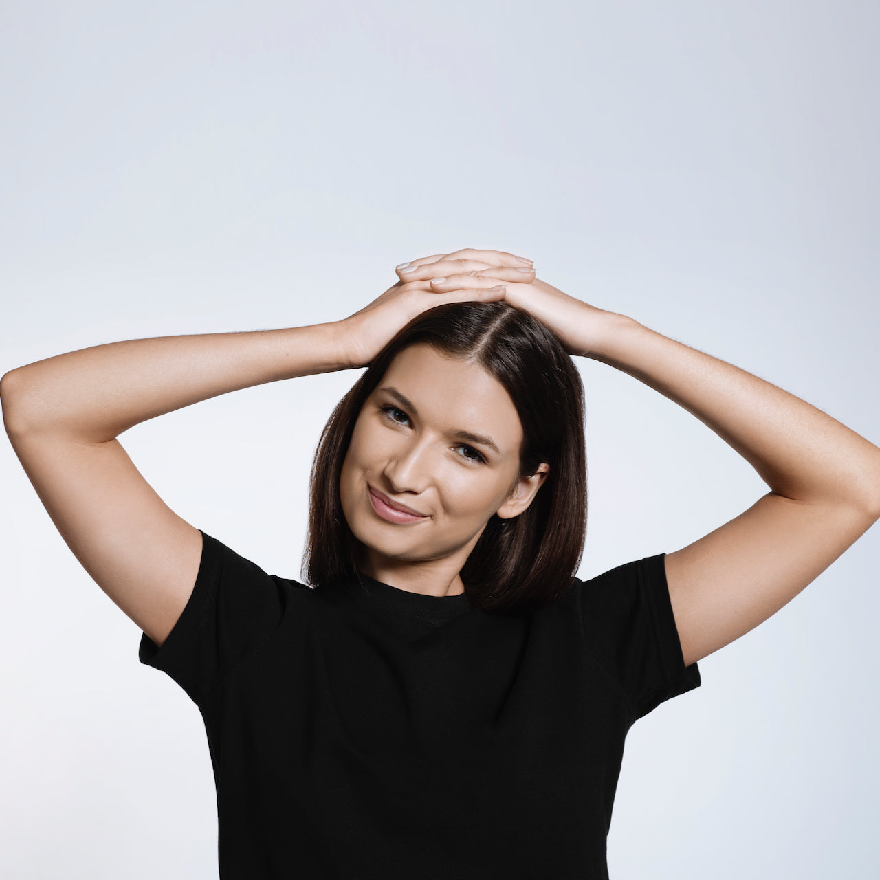 A woman smiling into the camera with her hands on her head, modelling a plain black short sleeve organic cotton t shirt.