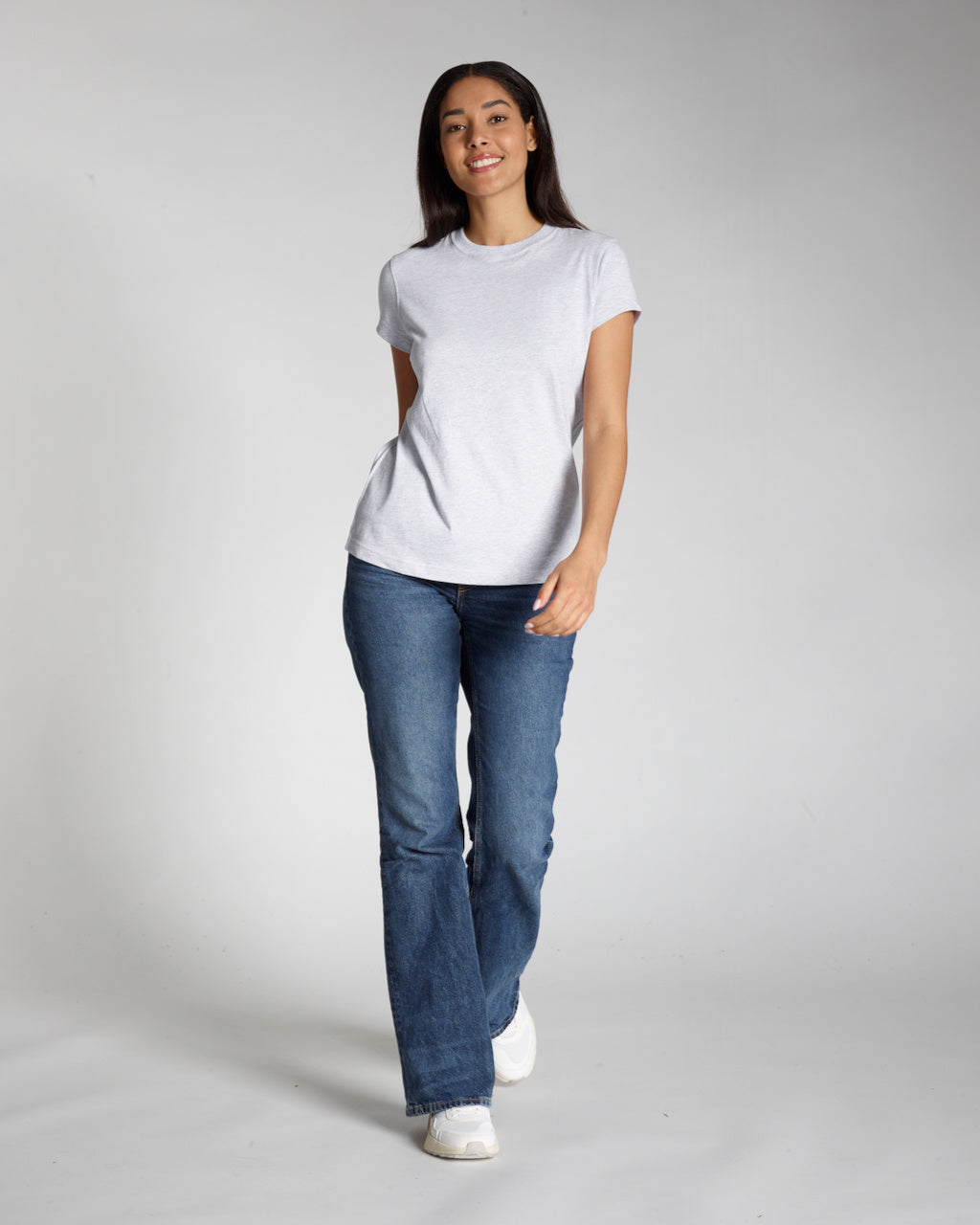 A full body photo of a woman smiling into the camera, modelling a plain grey, short sleeve organic cotton t shirt with a pair of jeans.
