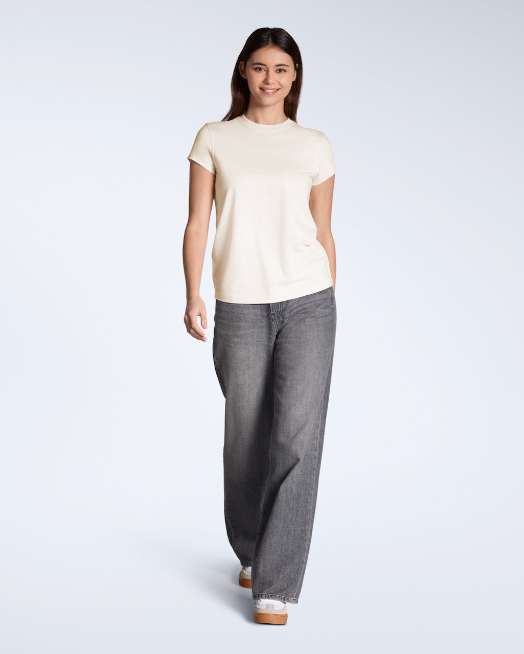 A full body photo of a woman smiling into the camera, modelling a plain natural coloured, short sleeve organic cotton t shirt with a pair of jeans.