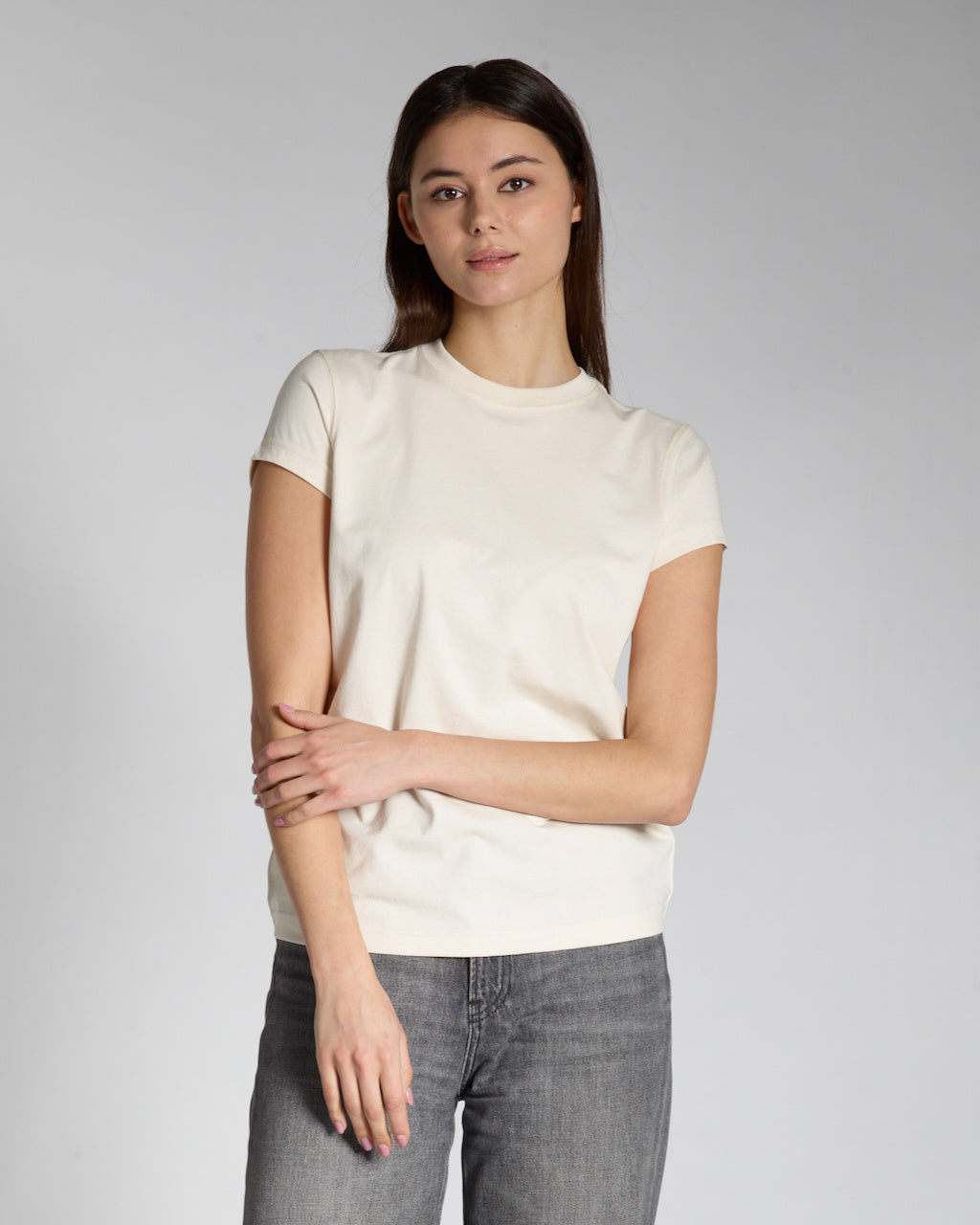 A woman looking into the camera touching her arm, modelling a plain natural coloured, short sleeve organic cotton t shirt.