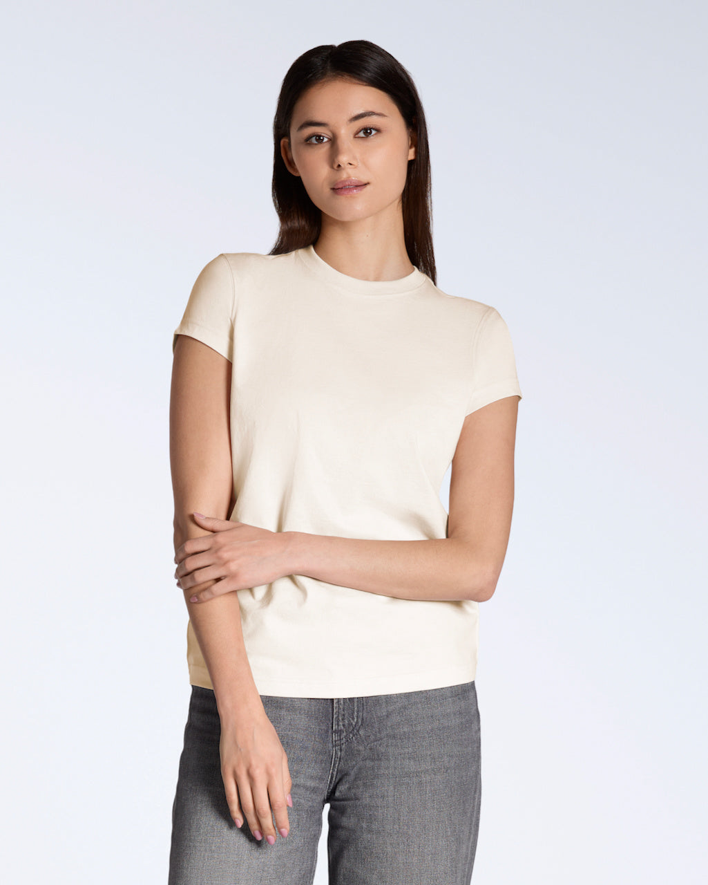 A woman looking into the camera touching her arm, modelling a plain natural coloured, short sleeve organic cotton t shirt.