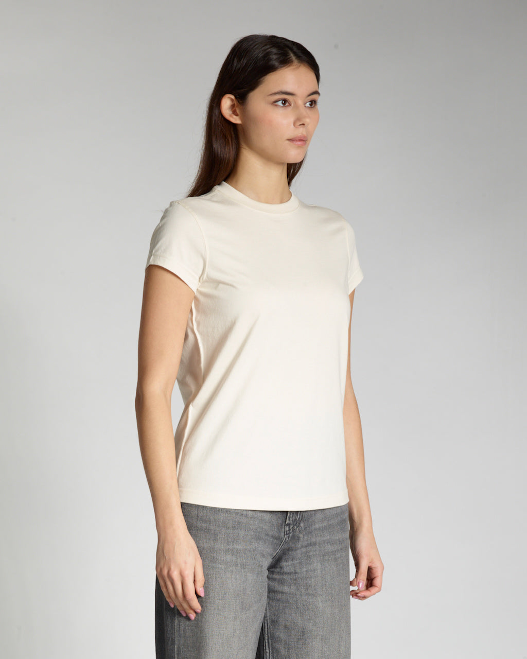 A woman at an angle with her arms by her sides, modelling a plain natural coloured, short sleeve organic cotton t shirt.