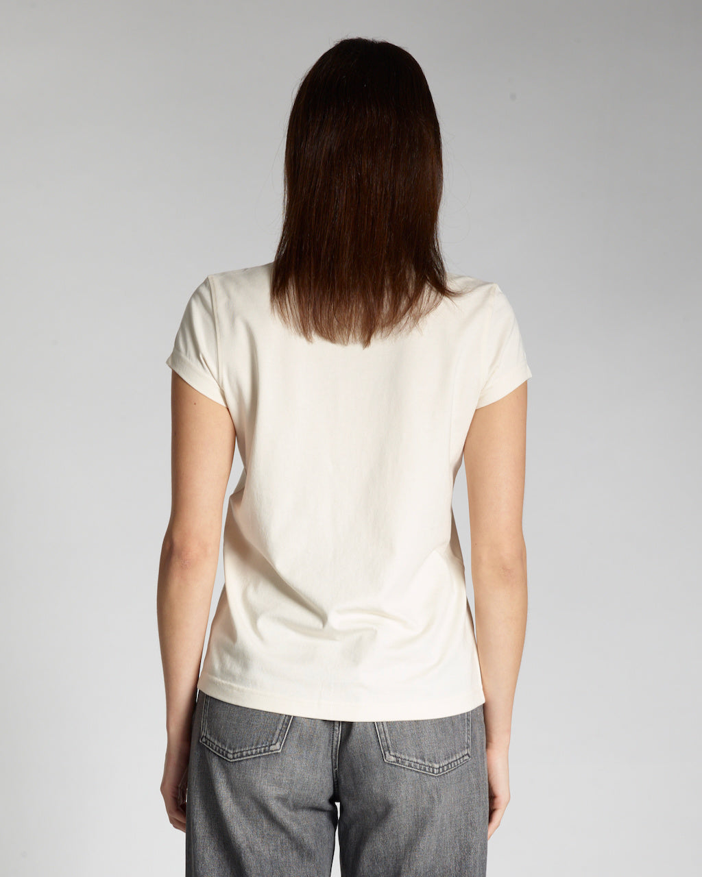 A woman facing away from the camera, modelling a plain natural coloured, short sleeve organic cotton t shirt.
