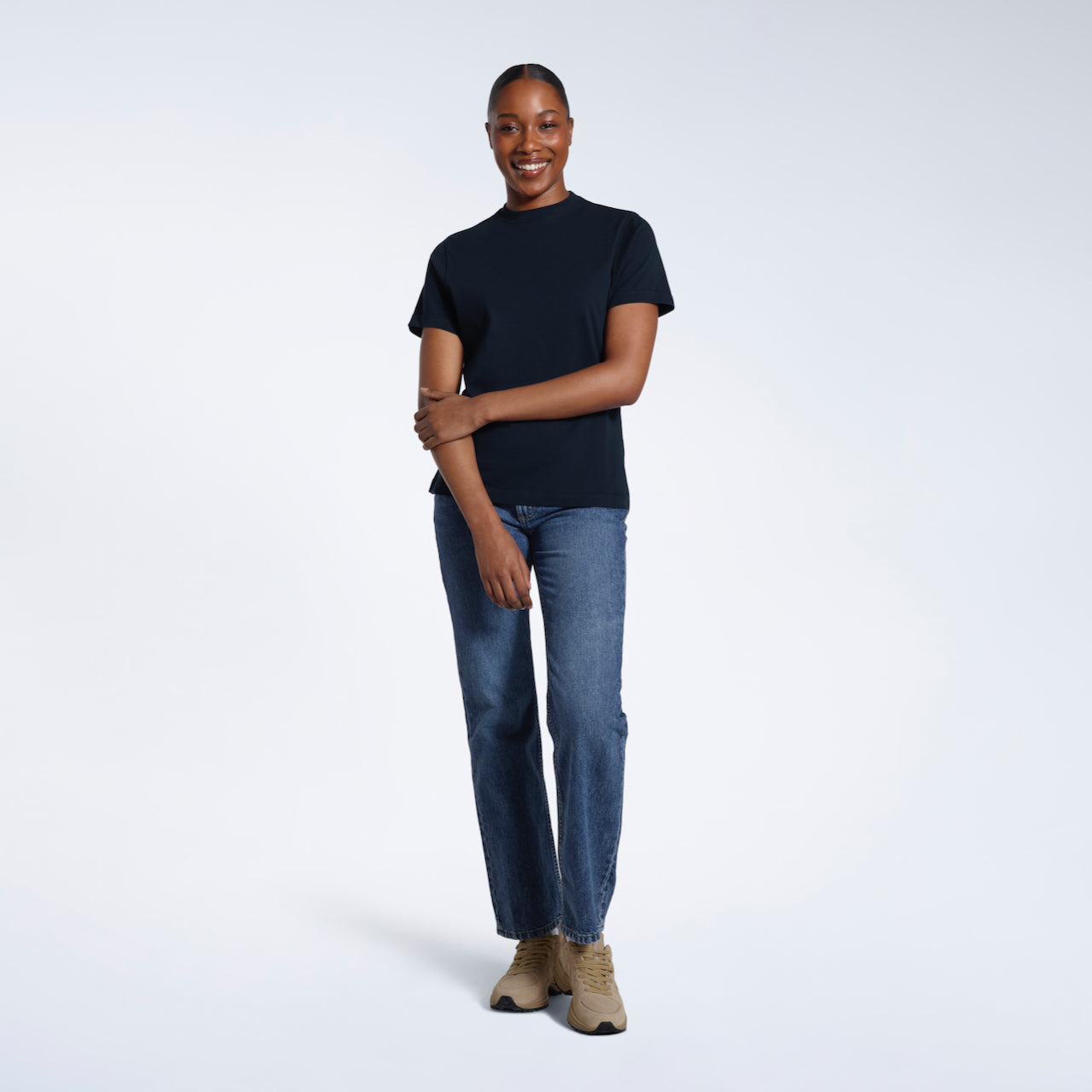 A full body photo of a woman smiling touching her arm, modelling a plain navy short sleeve organic cotton t shirt.