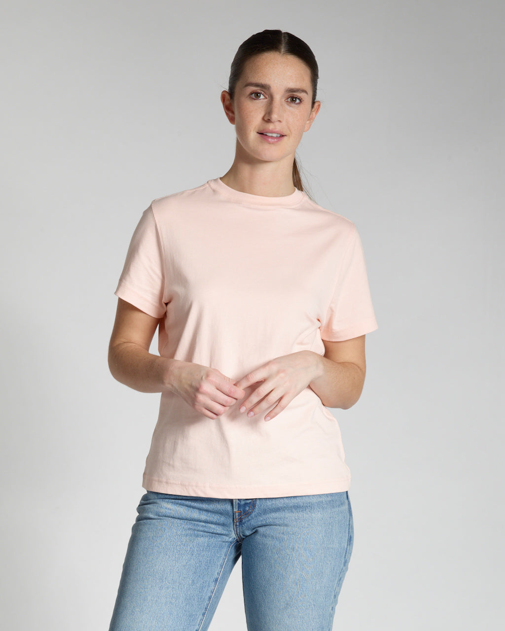 A woman looking into the camera touching her hand, modelling a plain pink, short sleeve organic cotton t shirt.