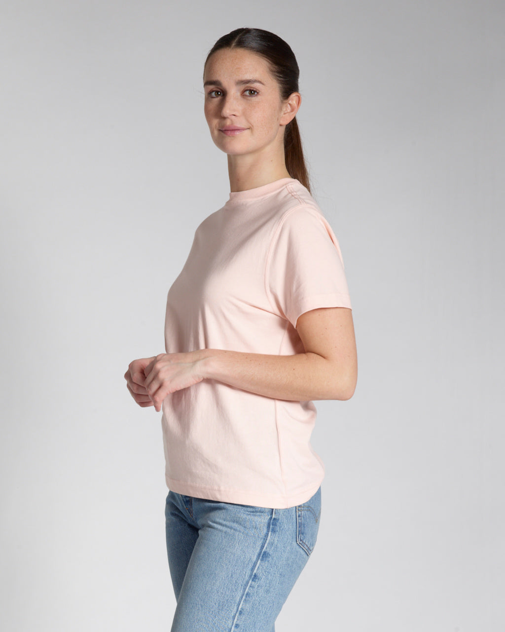 A woman at an angle smiling into the camera, modelling a plain pink, short sleeve organic cotton t shirt.