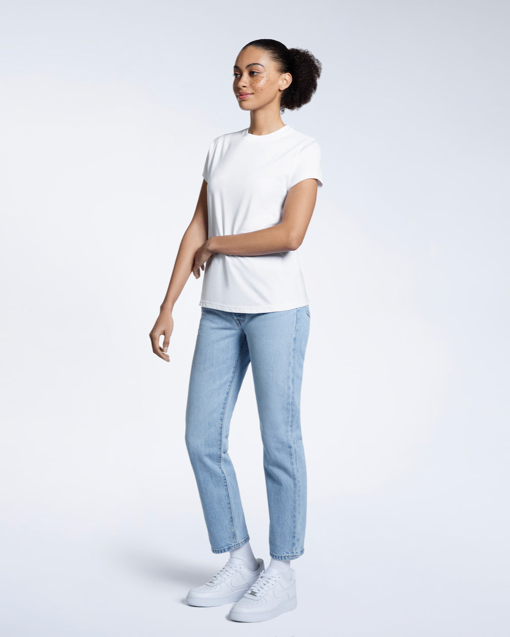 A full body photo of a woman touching her arm, modelling a plain white short sleeve organic cotton t shirt.