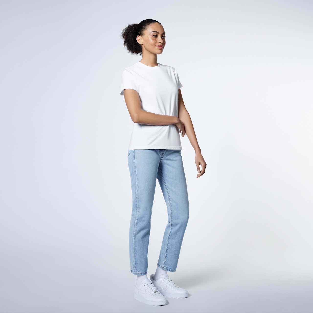 A full body photo of a woman touching her arm, modelling a plain white short sleeve organic cotton t shirt.