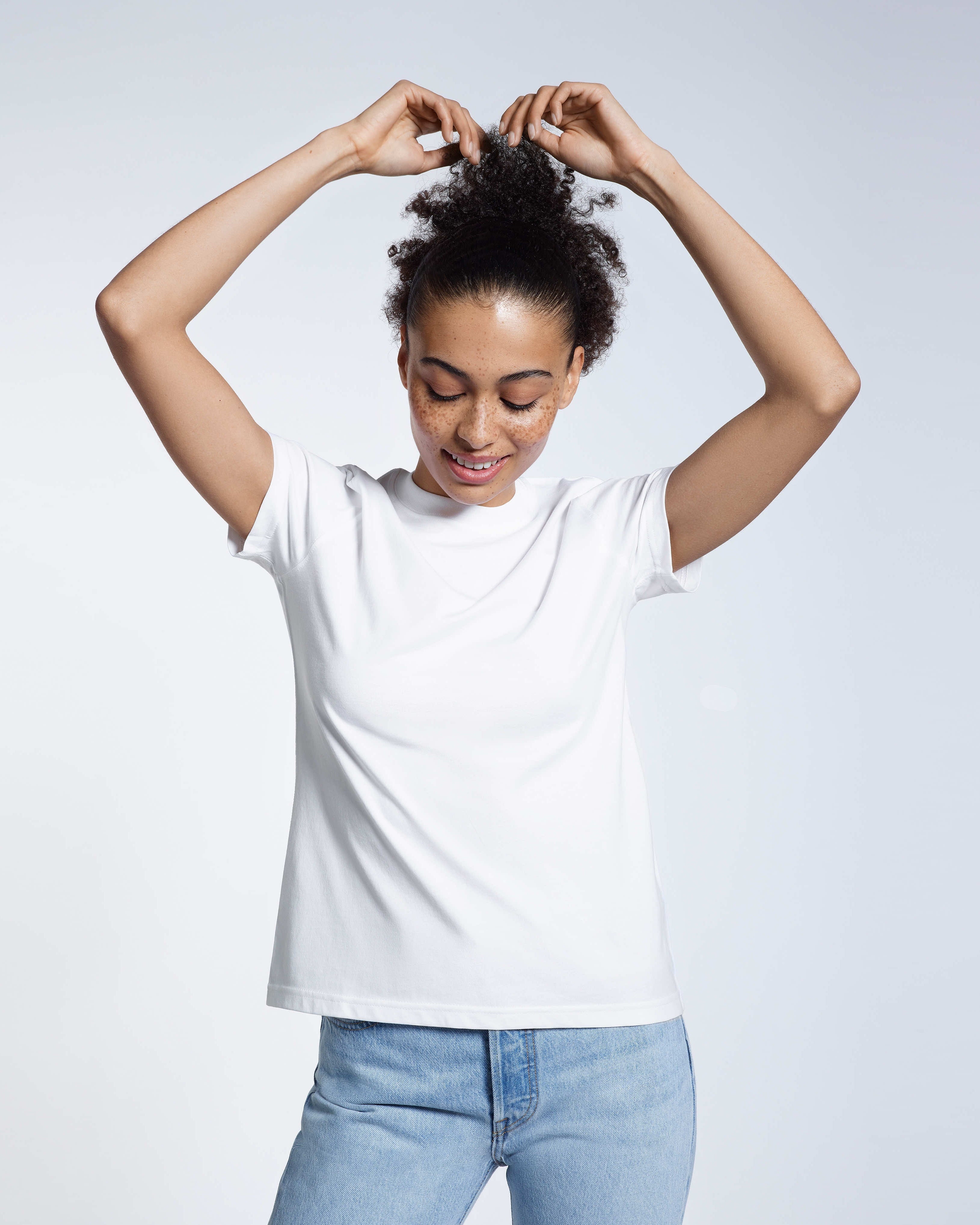 A woman with her arms above her head touching her hair, modelling a plain white short sleeve organic cotton t shirt.