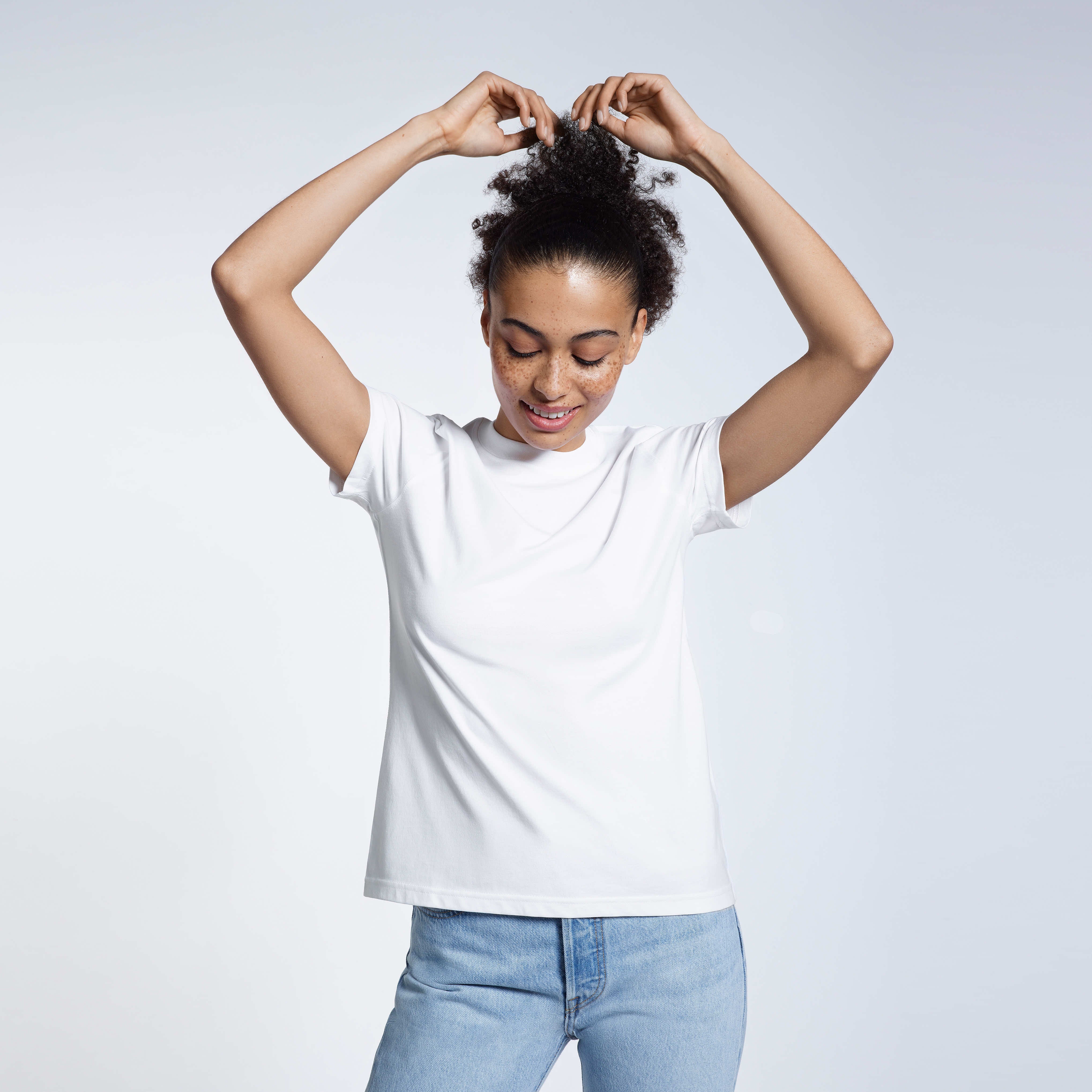 A woman with her arms above her head touching her hair, modelling a plain white short sleeve organic cotton t shirt.