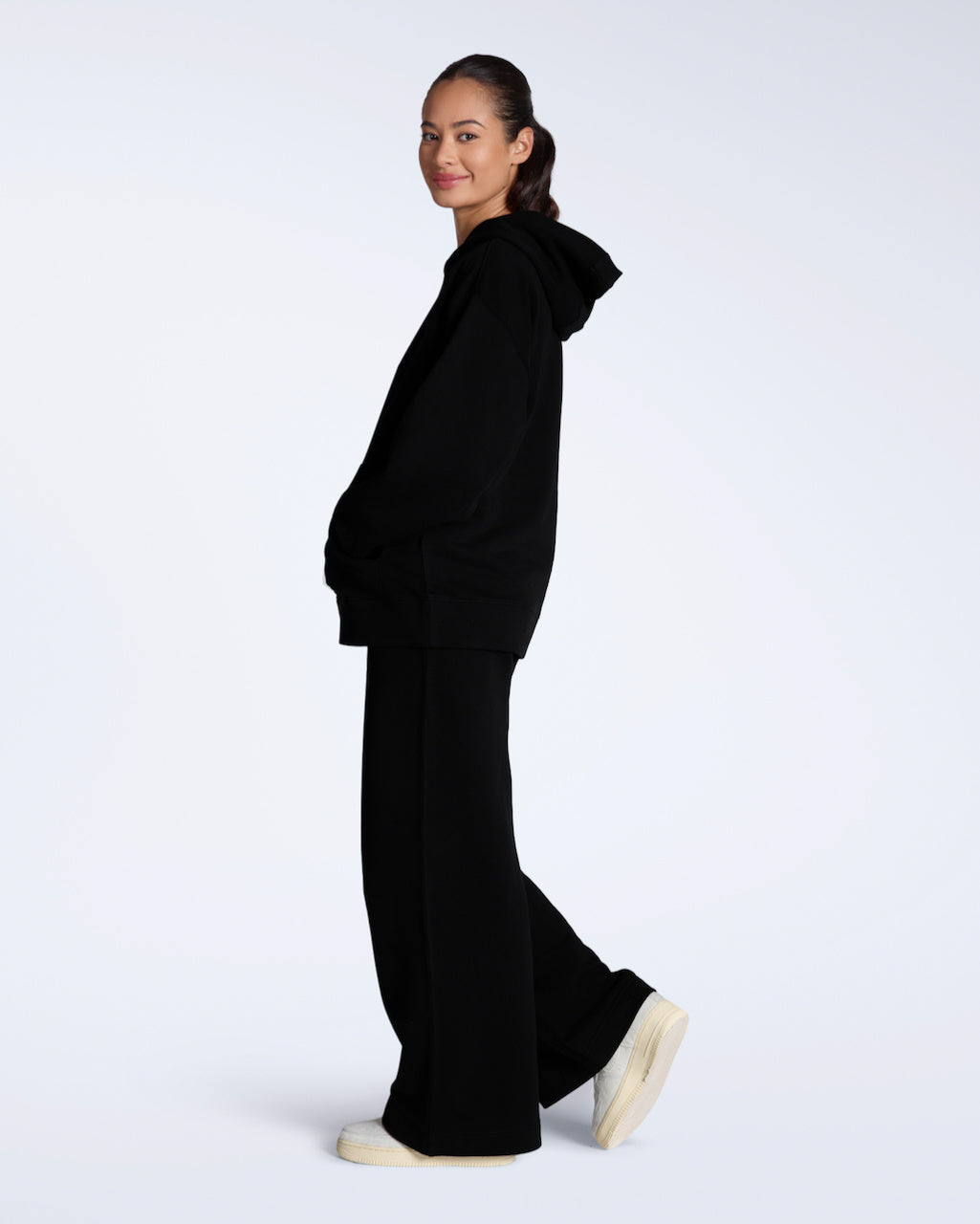 A woman facing side on looking back at the camera, modelling a black, organic cotton, oversized hoodie, with a pair of matching black, wide leg joggers.
