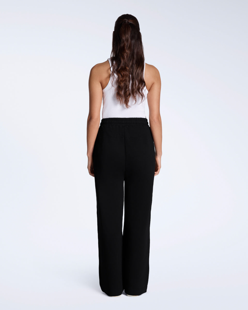 A woman facing away from the camera with her hands by her sides, modelling a pair of black, organic cotton, wide leg joggers.