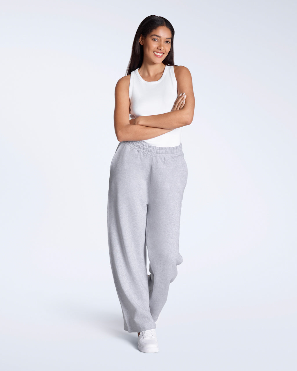 A woman with her arms crossed, smiling into the camera, modelling a white, organic cotton tank top with a pair of wide leg joggers.