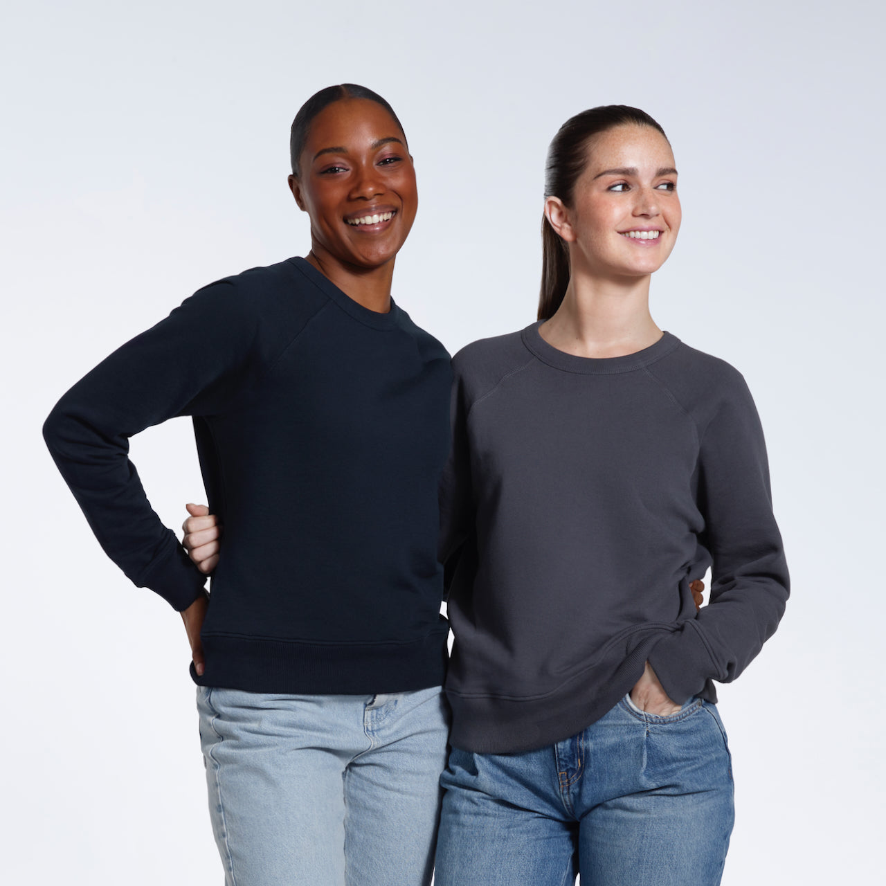 Two women with their arms round eachother smiling, modelling plain navy and dark grey organic cotton sweatshirts.