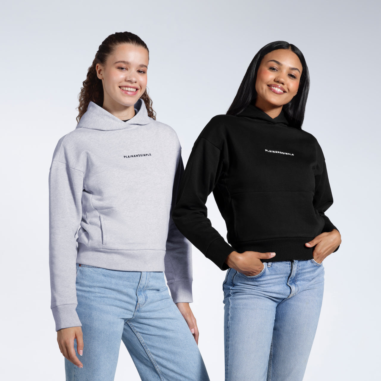 Two women looking at the camera smiling, modelling branded black and grey, heavyweight, organic cotton hoodies.