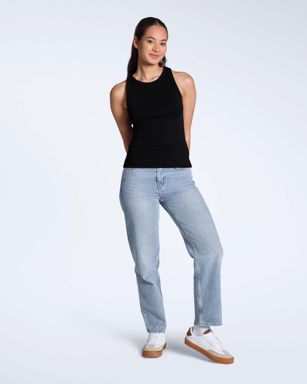 A full body photo of a woman smiling into the camera, modelling a plain black, organic cotton tank top with a pair of jeans.