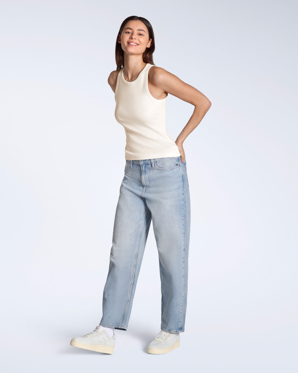 A full body photo of a woman smiling into the camera, modelling a plain natural coloured, organic cotton tank top with a pair of jeans.