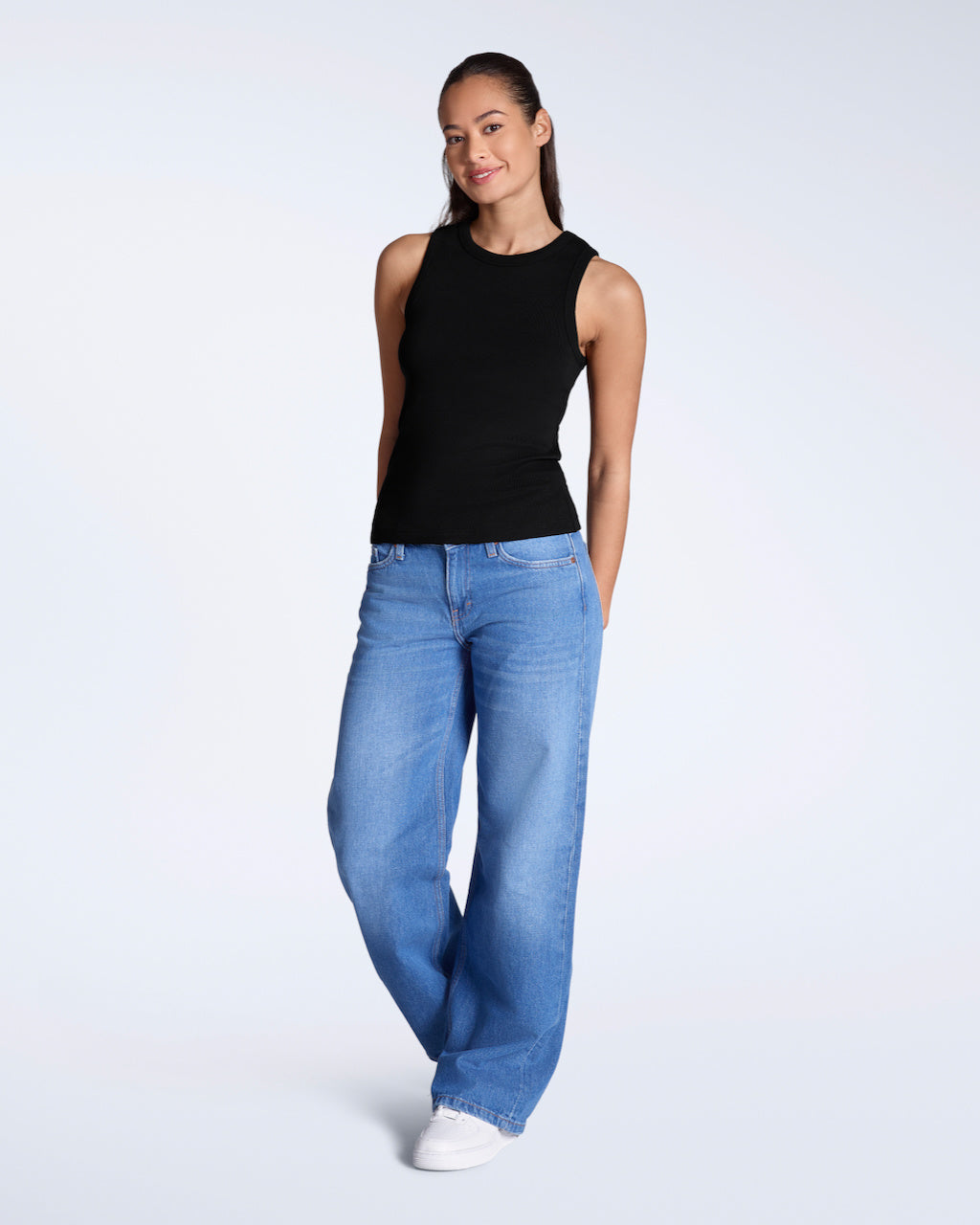 A full body photo of a woman smiling into the camera, modelling a plain black, organic cotton tank top with a pair of jeans.