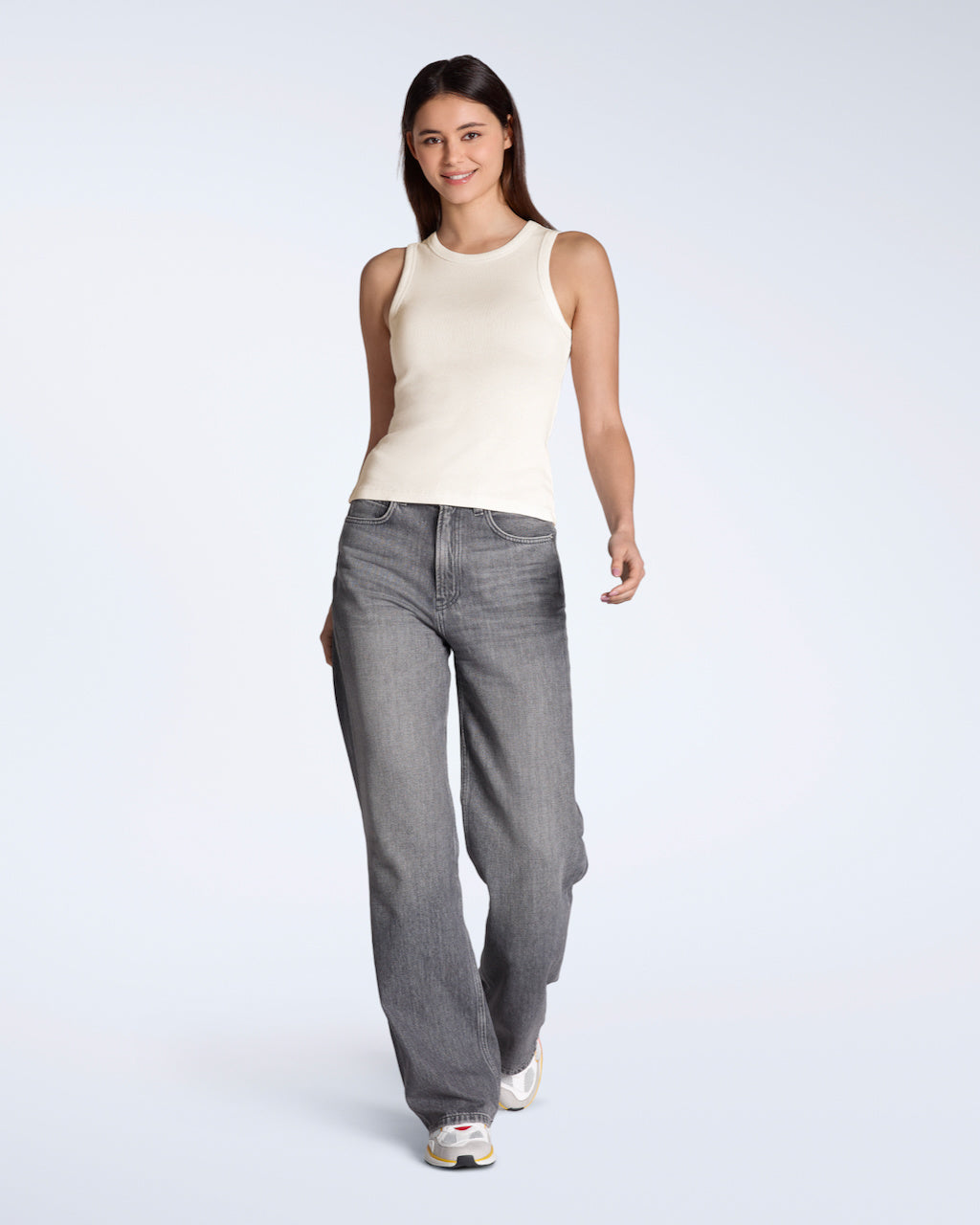 A full body photo of a woman smiling into the camera, modelling a plain natural coloured, organic cotton tank top with a pair of jeans.