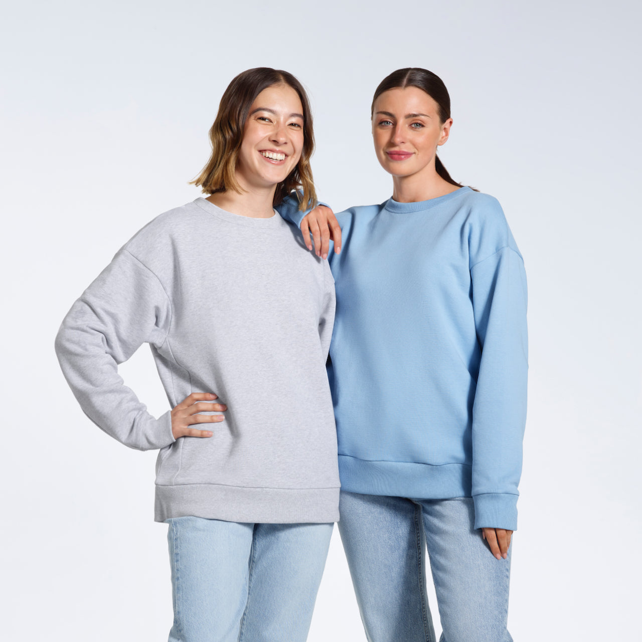 Two women smiling with their arms round eachother, modelling plain grey and light blue oversized, organic cotton sweatshirts.