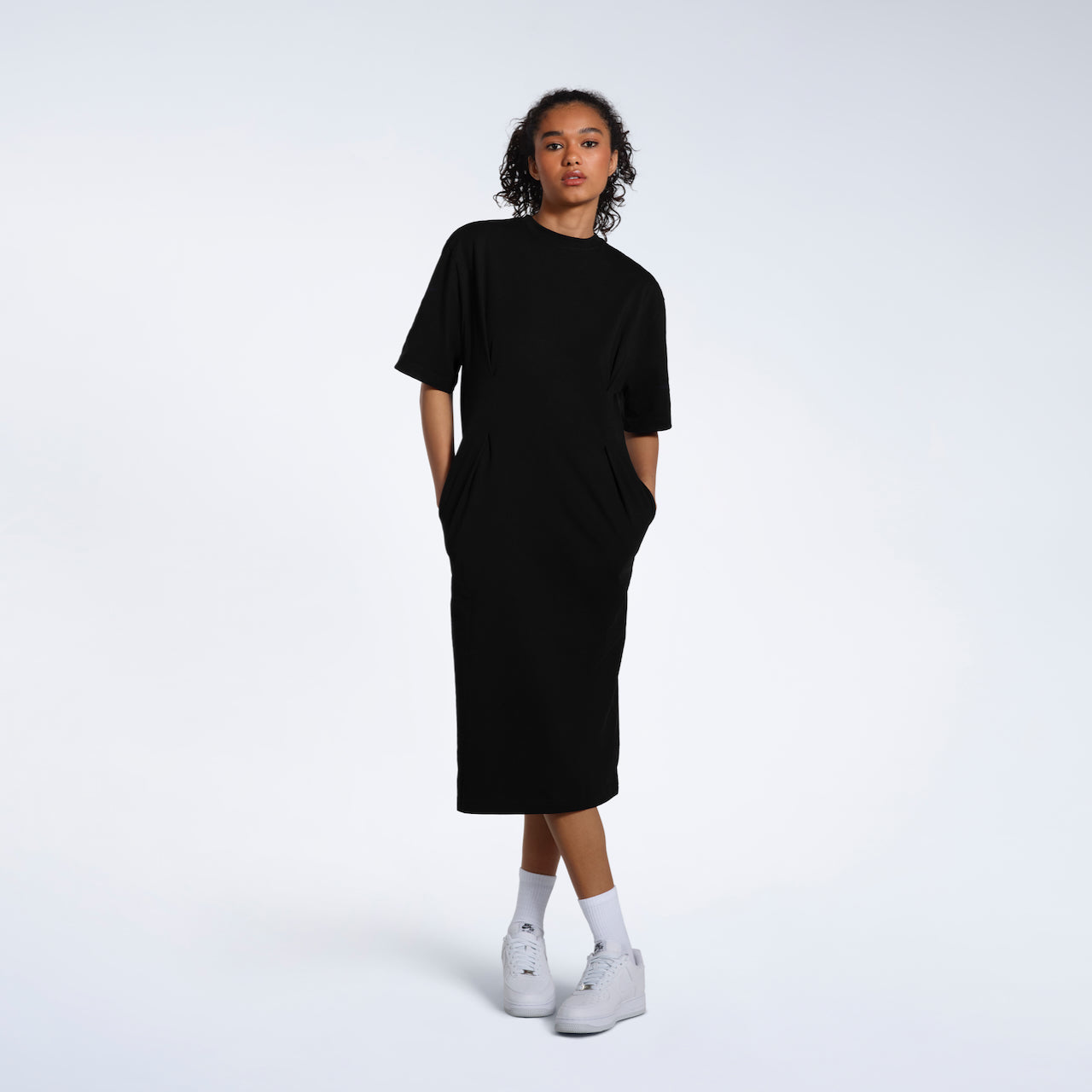 A women looking into the camera with her hands in her pockets, modelling a plain black, organic cotton midi t shirt dress.