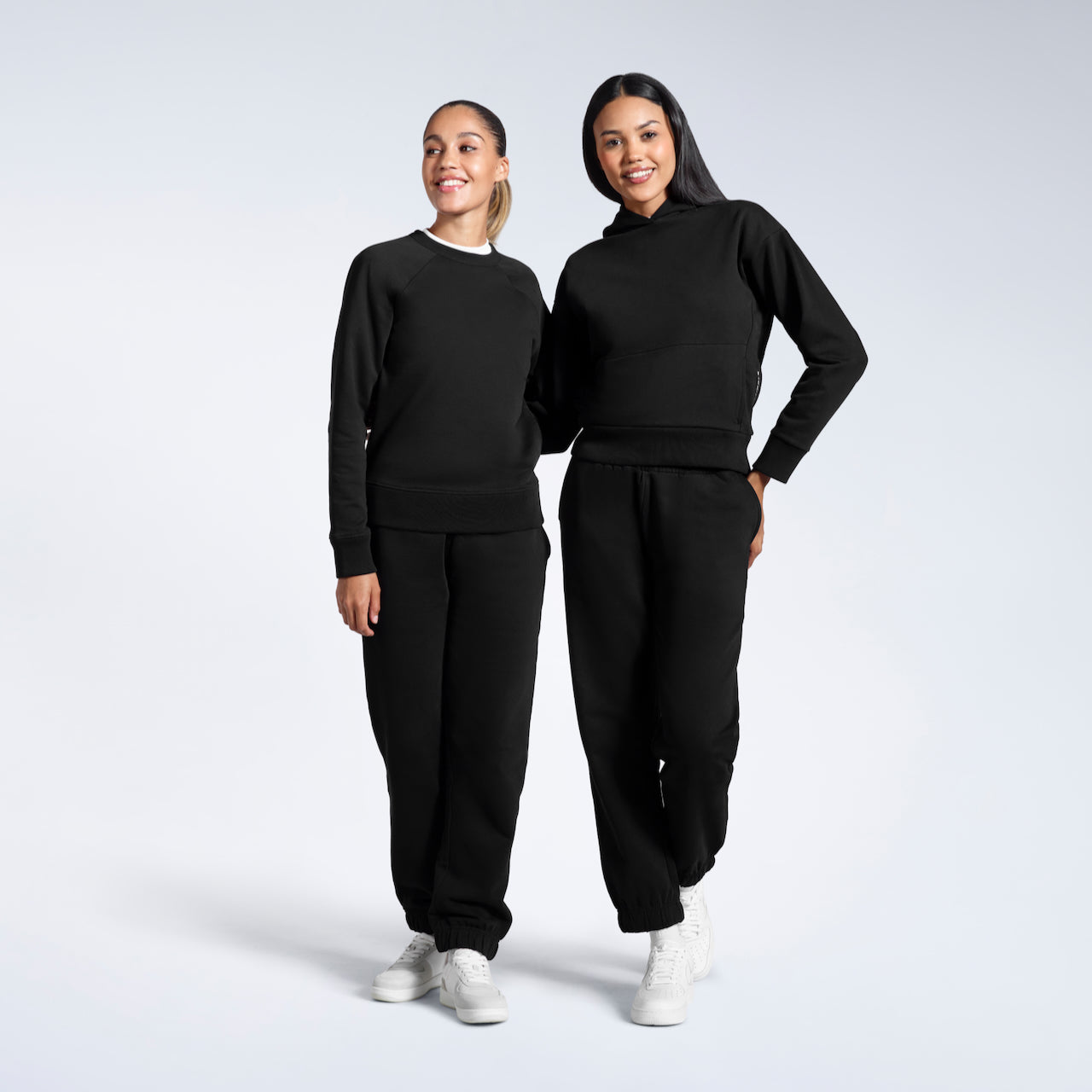 Two women with their arms round eachother smiling, modelling plain black, heavyweight, organic cotton tracksuit sets.