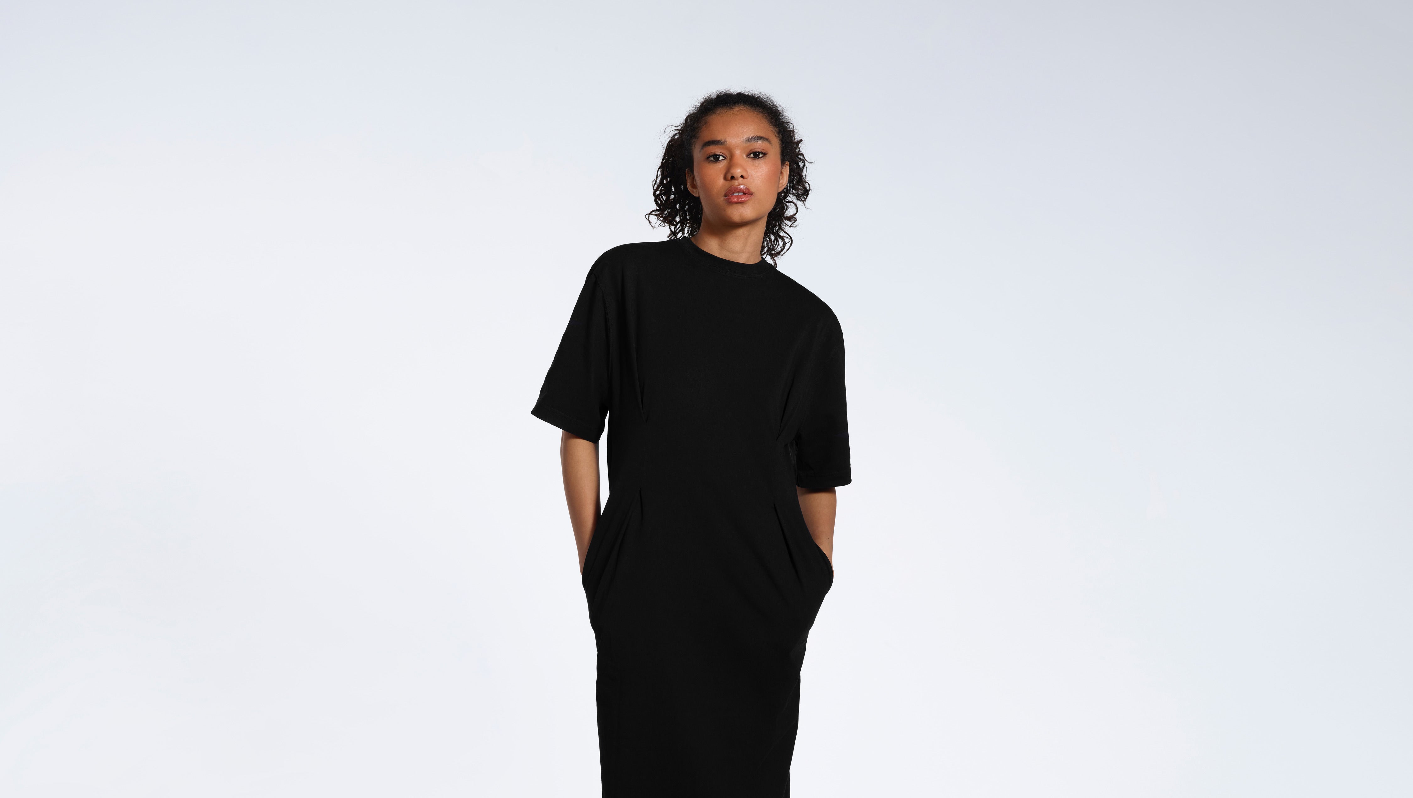 A woman looking into the camera with her hands in her pockets, modelling a plain black organic cotton t shirt dress.