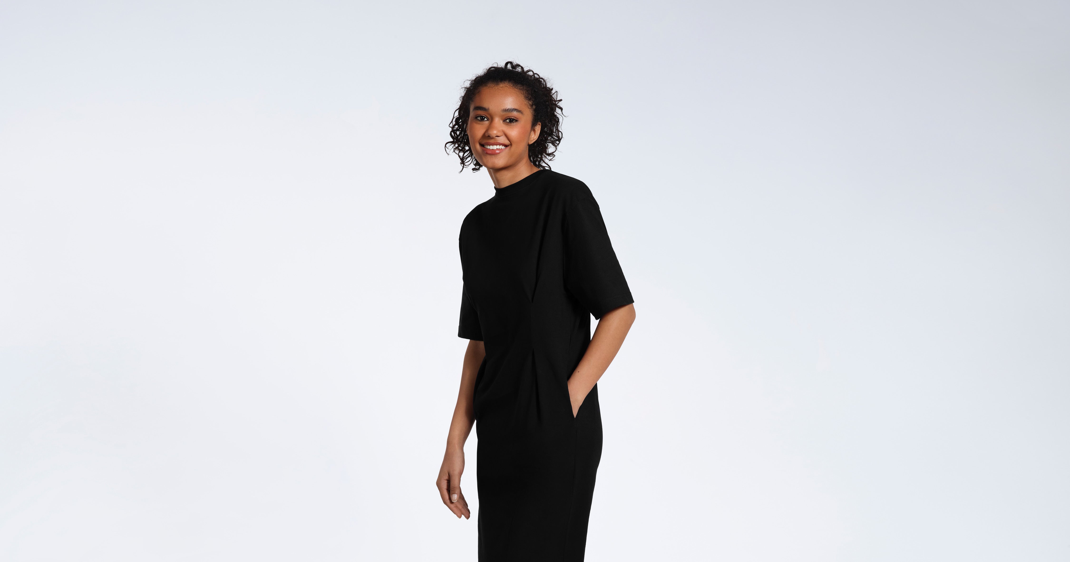 A woman looking into the camera smiling with her hand in her pocket, modelling a plain black organic cotton t shirt dress.