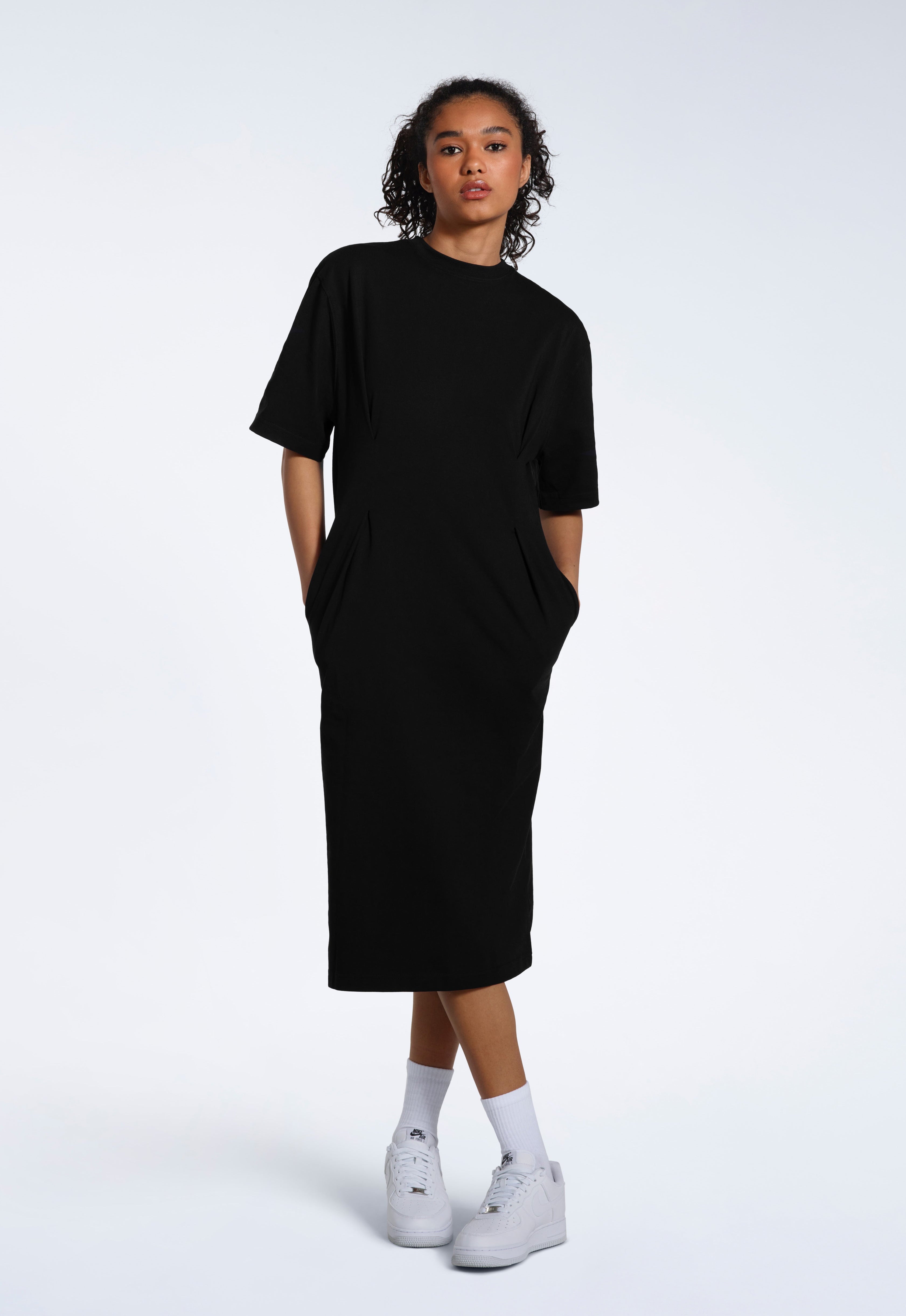 A woman looking into the camera with her hands in her pockets, modelling a plain black organic cotton t shirt dress.