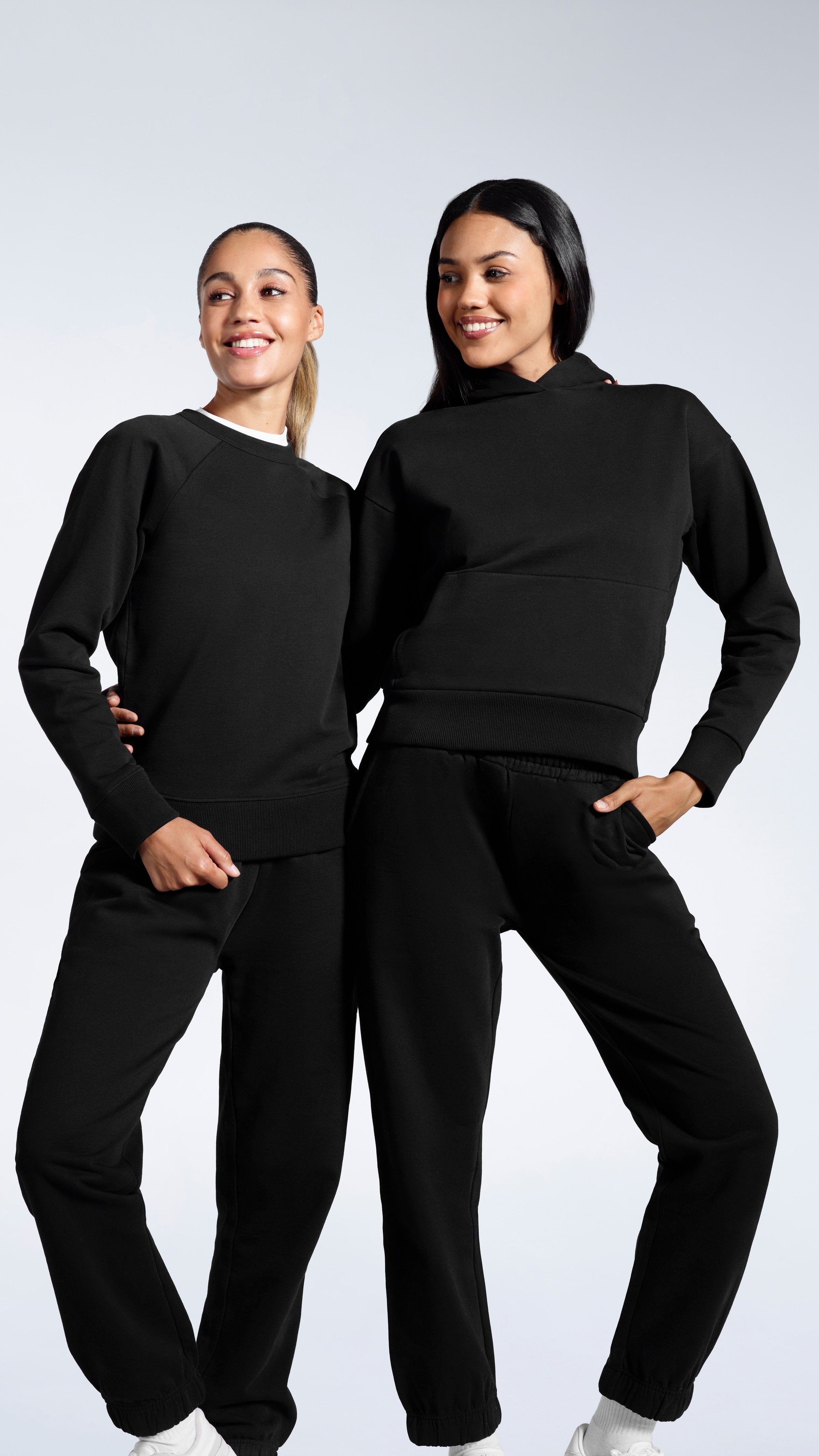 Two women in a plain black tracksuit set, smiling against a grey background.
