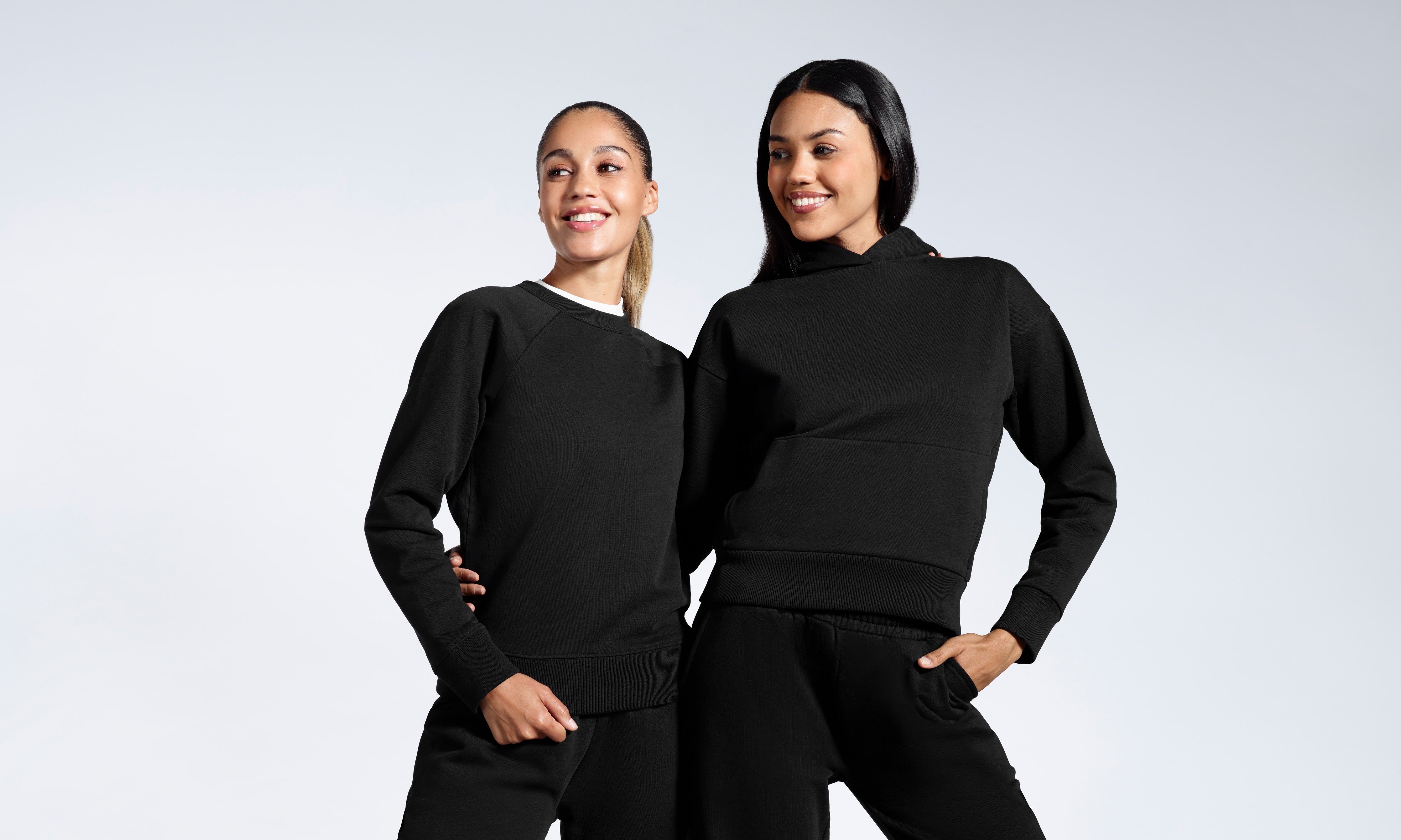 Two women with their arms round eachother smiling, modelling plain black, heavyweight, organic cotton tracksuit sets.